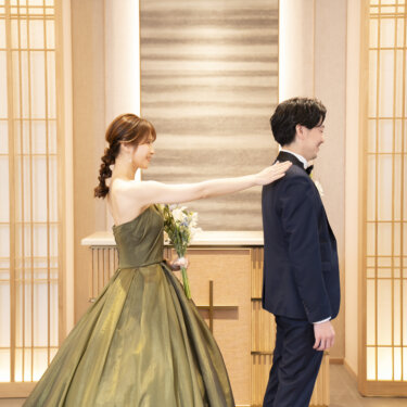 三つ編みヘアにカーキのドレスの花嫁が新郎の肩を後ろからたたく瞬間。スタジオフォトで人気のファーストミートの演出