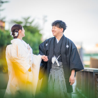 緑の木々を前ボケにして、笑顔で見つめ合う白無垢の花嫁と紋付袴の新郎。自然体の表情を写した和装前撮り