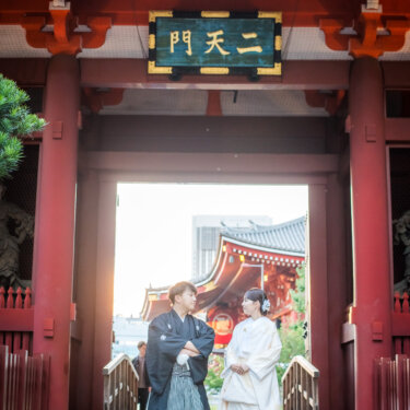 浅草寺の二天門の下で腕を組んで並ぶ紋付袴の新郎と白無垢の花嫁。門越しに朱色のお堂が見える和装前撮り