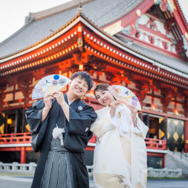 朱色のお堂の前で扇子を顔の横に掲げ、おどけた笑顔を見せる紋付袴の新郎と白無垢の花嫁の結婚写真