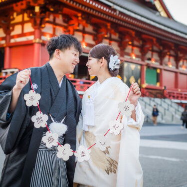 浅草寺本堂を背景に、花形のガーランドを持って見つめ合う紋付袴と白無垢の新郎新婦。和装前撮りのアイテム使い