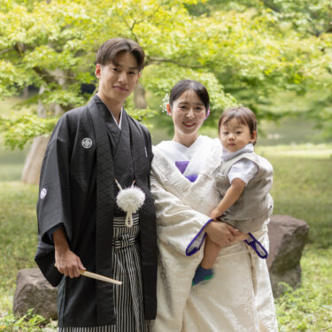新緑の庭で男の子を抱いた白無垢の花嫁と、扇子を持つ紋付袴の新郎がそろって笑顔を見せる家族写真の和装前撮り