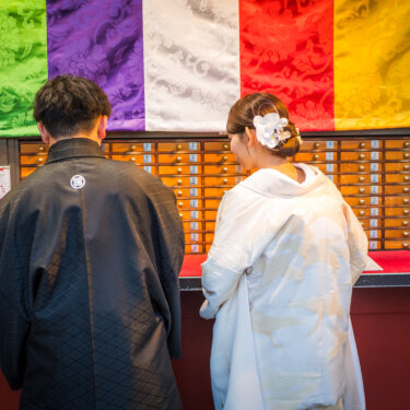 色鮮やかな五色の幕の下、おみくじの引き出しの前に並ぶ紋付袴の新郎と白無垢の花嫁の後ろ姿の和装前撮り