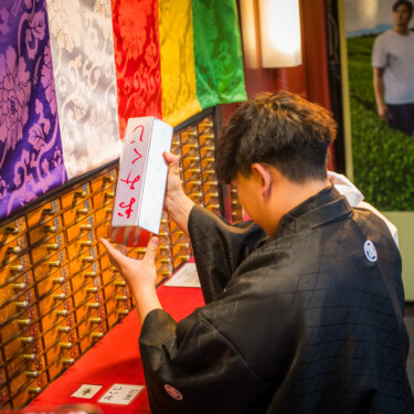 浅草寺のおみくじ所で六角形のおみくじ筒を振る紋付袴の新郎。浅草ならではの体験を写した和装フォトウェディング