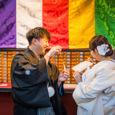 五色の幕とおみくじの棚の前で、引いたおみくじを笑顔で開く紋付袴の新郎と白無垢の花嫁の前撮り写真