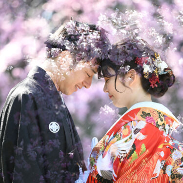 枝垂れ桜の枝越しにおでこを寄せ合う紋付袴の新郎と、鶴柄の赤い色打掛の花嫁。桜で撮る和装前撮りで人気のおでこ合わせポーズ