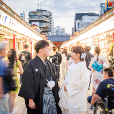 人通りの多い商店街の参道の真ん中に立つ紋付袴の新郎と白無垢の花嫁。にぎわう街並みで撮る和装前撮り