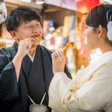 仲見世通りの店先で、串に刺さった団子を頬張る紋付袴の新郎と白無垢の花嫁。浅草の食べ歩き和装前撮り