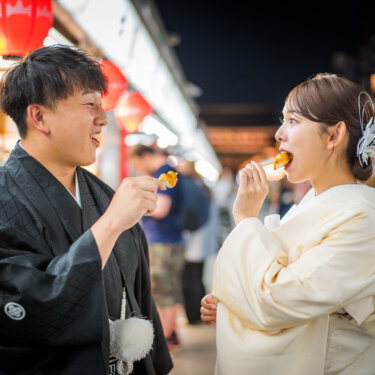 夜の仲見世通りで提灯の明かりの下、団子を食べながら笑う紋付袴の新郎と白無垢の花嫁の和装フォトウェディング
