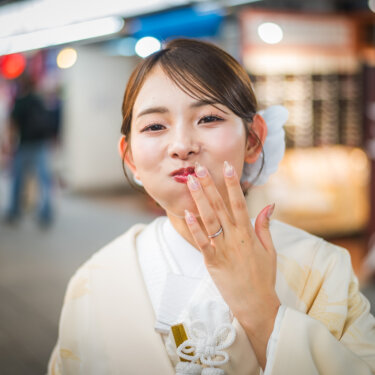 にぎやかな参道を背景に、口元に手を添えて照れ笑いする白無垢の花嫁のアップ。自然な表情の和装前撮り