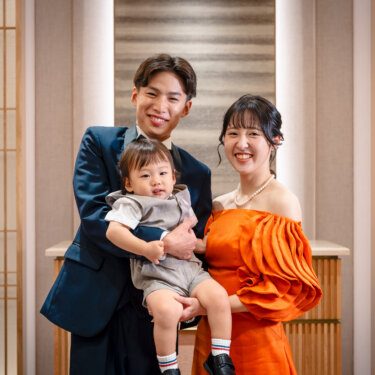 和モダンなチャペルで、息子を抱く新郎とオレンジのカラードレスの花嫁が笑顔で並ぶ家族写真。子どもと一緒に撮るフォトウェディング