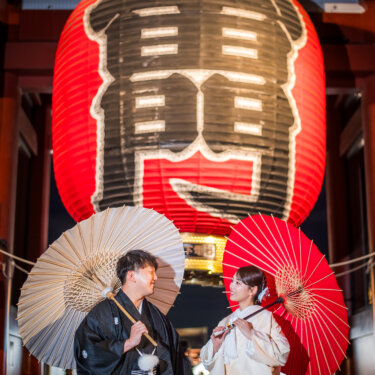 浅草寺の大提灯の下で、白と赤の和傘をさして見つめ合う紋付袴の新郎と白無垢の花嫁。浅草の夜の和装前撮り