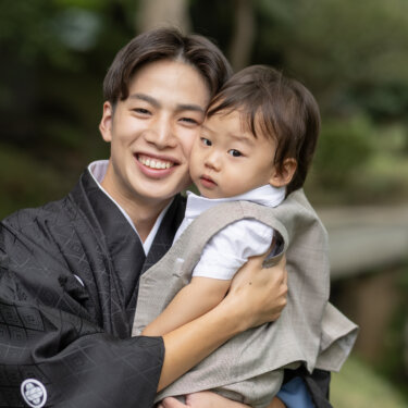 木立の中で男の子を抱き寄せ、ほおを寄せて笑う紋付袴の新郎。和装前撮りで残す親子の家族写真