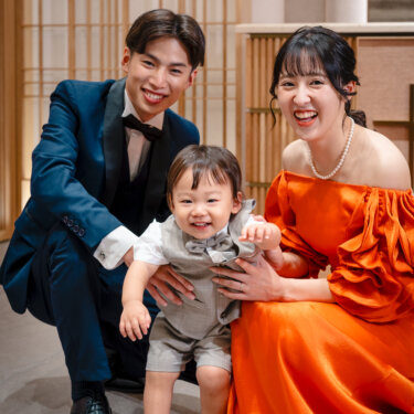 床にしゃがんで小さな息子を支え、満面の笑みを見せるタキシードの新郎とカラードレスの花嫁。ベストに蝶ネクタイの男の子と撮る家族写真の前撮り