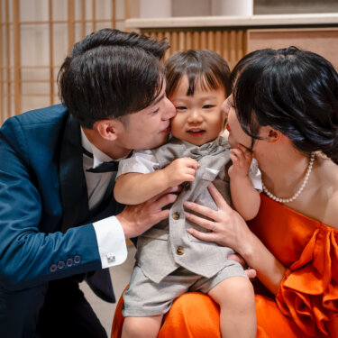 ベスト姿の息子の頬に両側からキスをするタキシードの新郎とカラードレスの花嫁。家族で撮るフォトウェディングで人気のほっこりポーズ