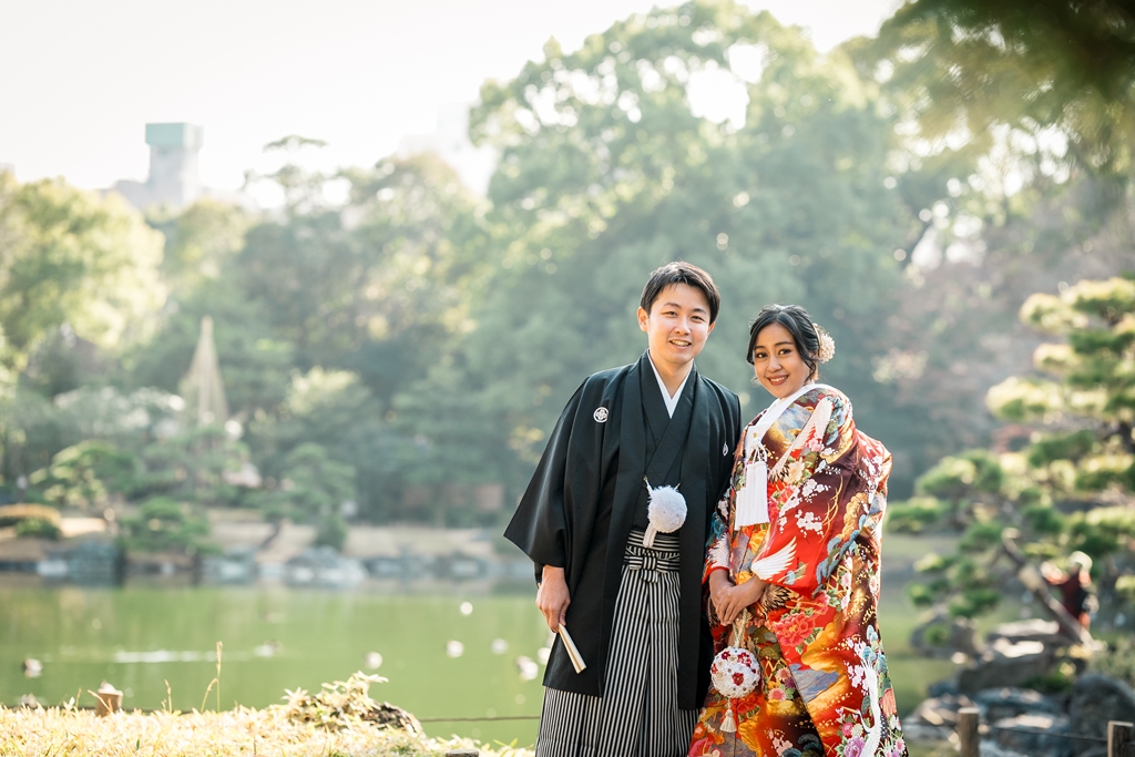 清澄庭園の池を背景に笑顔で並ぶ赤い色打掛の花嫁と黒紋付袴の新郎。東京の日本庭園での和装フォトウェディング
