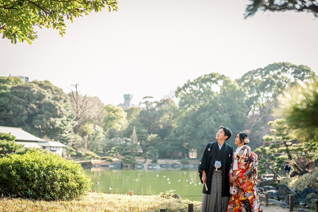 清澄庭園の大泉水のほとりに立つ赤い色打掛の花嫁と黒紋付袴の新郎。和装前撮りで人気の東京の日本庭園
