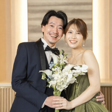 白いカラーのブーケを持つカーキのドレスの花嫁と頬を寄せて笑うネイビーのタキシードの新郎。東京駅店のスタジオで撮る洋装前撮り