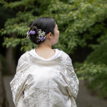 もみじの緑を背に、紫の花と白いかすみ草の髪飾りを見せる白無垢の花嫁の後ろ姿。和装前撮りのヘアアレンジ