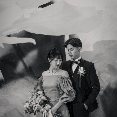 花びらを描いた大きなアートを背に寄り添う、タキシードの新郎とパフスリーブのカラードレスの花嫁。モノクロで仕上げたクラシカルな結婚写真