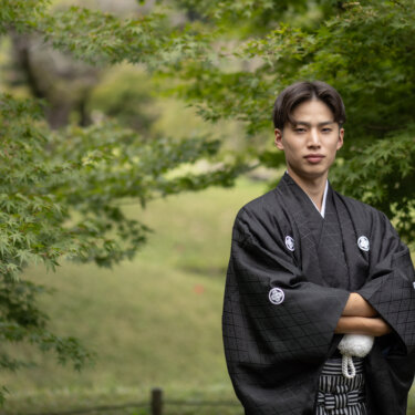 新緑のもみじの下で腕を組んでカメラを見つめる黒紋付袴の新郎。小石川後楽園で撮る和装前撮りの新郎ショット