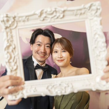 花柄のアートウォールの前で白い額縁から顔をのぞかせて微笑むタキシードの新郎とカラードレスの花嫁。額縁を使ったスタジオフォトの楽しい一枚