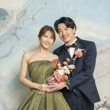 淡い色のアートウォールを背に、頬を寄せ合って微笑むタキシードの新郎とカーキのカラードレスの花嫁。スタジオフォトの定番ポーズ