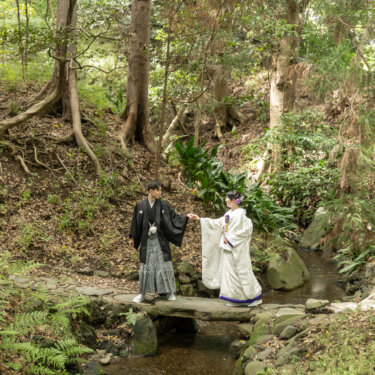 小石川後楽園の木々に囲まれた小川の石橋で、手を取り合う紋付袴の新郎と白無垢の花嫁。緑豊かな和装前撮り
