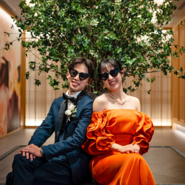 グリーンの大木の前に並んで座り、ハート形のサングラスで笑うタキシードの新郎とカラードレスの花嫁。小物で遊び心を添えたフォトウェディング