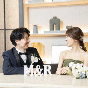 イニシャルのオブジェと白い花を置いたカウンターで見つめ合うタキシードの新郎と三つ編みヘアの花嫁。東京駅店のラウンジで撮る洋装前撮り