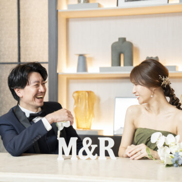 カウンターでイニシャルのオブジェと花を前に笑い合うタキシードの新郎とカーキのカラードレスの花嫁。リラックスした雰囲気のスタジオフォト