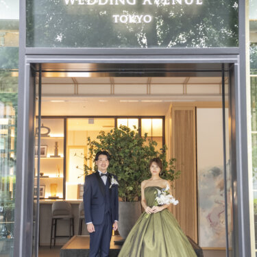ガラスの入口の前に立つ、ネイビーのタキシードの新郎とトレーンを広げたカーキのカラードレスの花嫁。東京駅店で撮る洋装前撮り