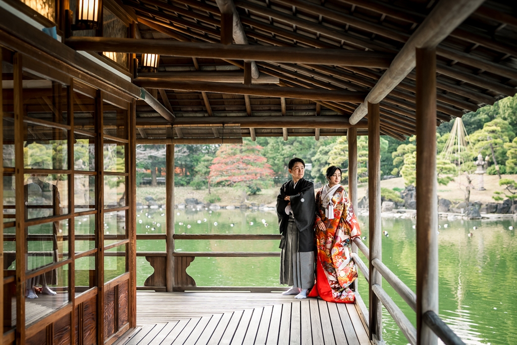 清澄庭園の涼亭の縁側に並んで立つ赤い色打掛の花嫁と紋付袴の新郎。池と庭園を背景にした和装前撮り