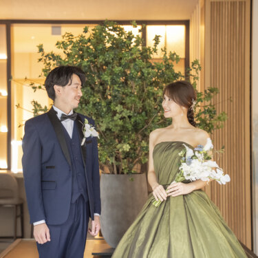 観葉植物と写真が飾られたラウンジで向かい合う、タキシードの新郎と白いブーケを持つオリーブグリーンのドレスの花嫁。東京駅店のラウンジでのスタジオフォト