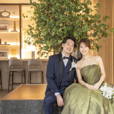 ラウンジのソファに並んで座って微笑む、ネイビーのタキシードの新郎とカーキのプリンセスラインのドレスの花嫁。東京駅店で撮る洋装前撮り