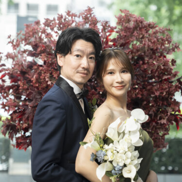赤い葉の植え込みを背に、白いカラーのブーケを持つカーキのドレスの花嫁を抱き寄せるネイビーのタキシードの新郎。東京駅店の屋外で撮る洋装前撮り