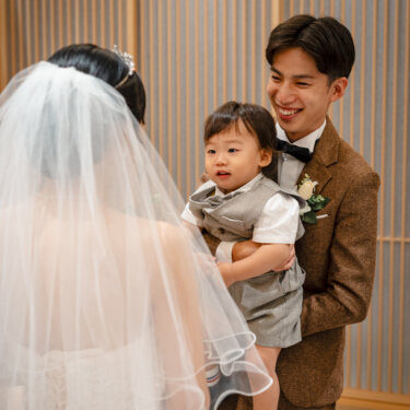 ブラウンのツイードスーツの新郎に抱かれた息子が、ベール姿の花嫁を見つめるワンシーン。花嫁の後ろ姿越しに撮った子連れのフォトウェディング