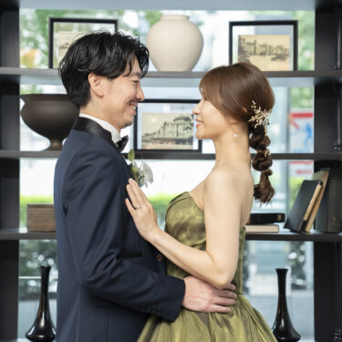 本棚と小物が並ぶラウンジで腰を抱き寄せて見つめ合うタキシードの新郎と三つ編みヘアの花嫁。東京駅店のラウンジで撮るおしゃれな洋装前撮り
