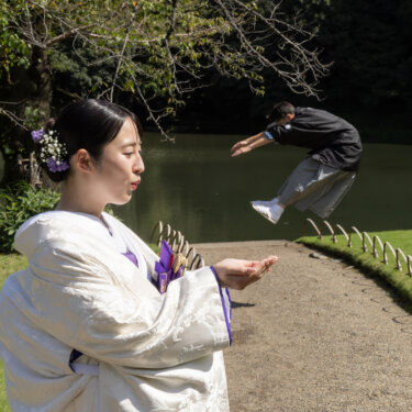 小石川後楽園の池のほとりで、遠くでジャンプする新郎を手のひらに乗せるように見せる白無垢の花嫁。遠近法を使った和装前撮りのポーズ