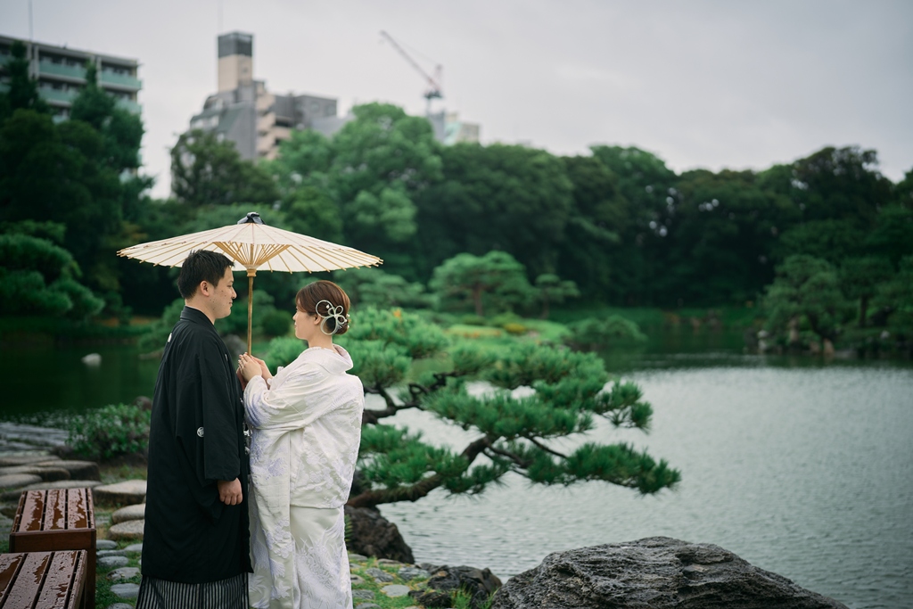 緑が深まる夏の清澄庭園で、和傘を差して池を眺める白無垢の花嫁と紋付袴の新郎