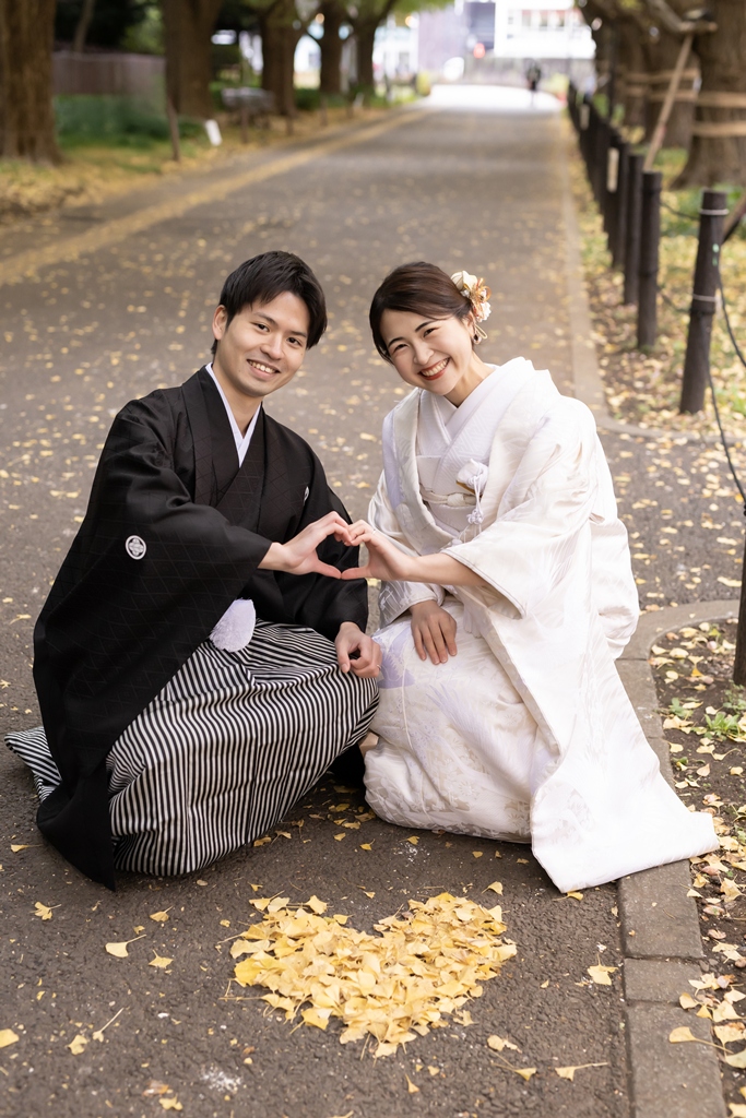 外苑いちょう並木の落ち葉で作ったハートの前に座り、手でハートを作る白無垢の花嫁と紋付袴の新郎
