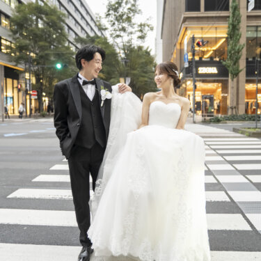 ビル街の横断歩道をドレスの裾を持ち上げて歩く、タキシードの新郎とウェディングドレスの花嫁。都会の街並みで撮る洋装前撮り