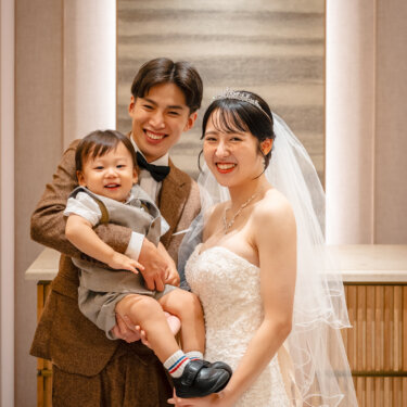 和モダンなチャペルで、息子を抱くブラウンのツイードスーツの新郎とビスチェのウェディングドレスの花嫁が笑顔で並ぶ家族写真の前撮り