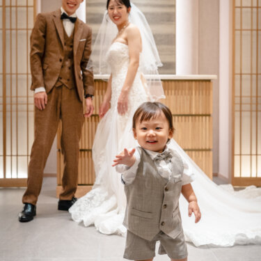 長いトレーンのウェディングドレスの花嫁と新郎を背に、カメラへ手を伸ばして歩いてくる息子。家族の自然な表情を残すフォトウェディング