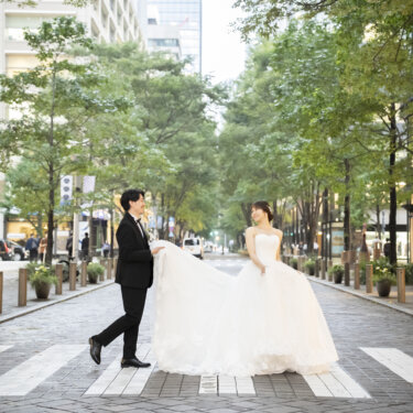 並木道の石畳の横断歩道で、ウェディングドレスの長い裾を広げて歩く新郎新婦。丸の内仲通りで撮るロケーションフォト