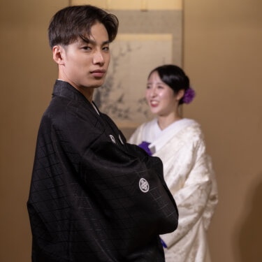 振り返ってカメラを見つめる紋付袴の新郎と、後ろでほほえむ白無垢の花嫁。和室で撮るクールな和装フォトウェディング