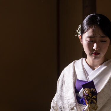 暗い和室で光を受けて目を伏せる、紫の小物を合わせた白無垢の花嫁のアップ。陰影が美しい和装前撮り