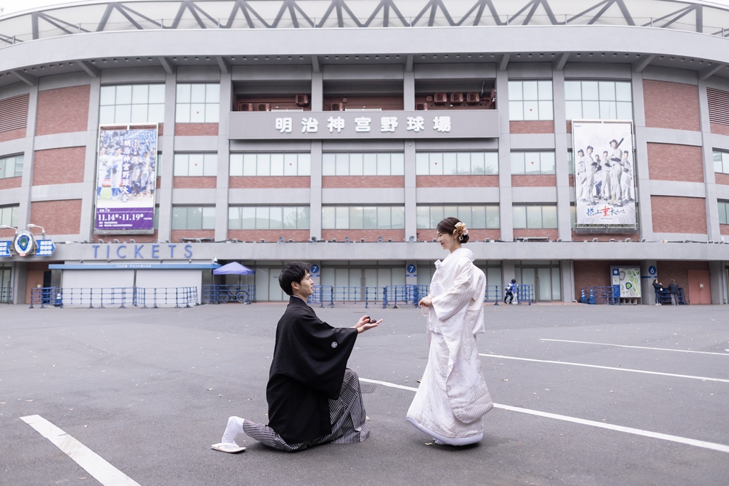 神宮球場の前で、ひざまずいてプロポーズを再現する紋付袴の新郎と白無垢の花嫁