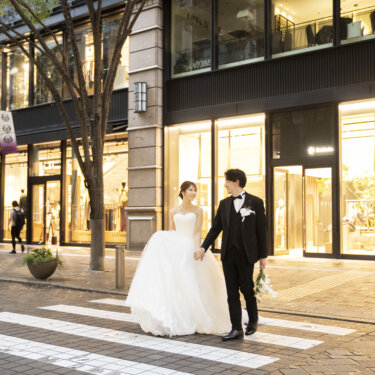 明かりの灯るショーウィンドウの前の横断歩道で、手をつないで歩く新郎新婦。夕方の街並みで撮るフォトウェディング