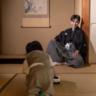 掛け軸の和室で畳にしゃがむ紋付袴の新郎と、はいはいで向かう男の子。家族で残す和装前撮りの自然な瞬間